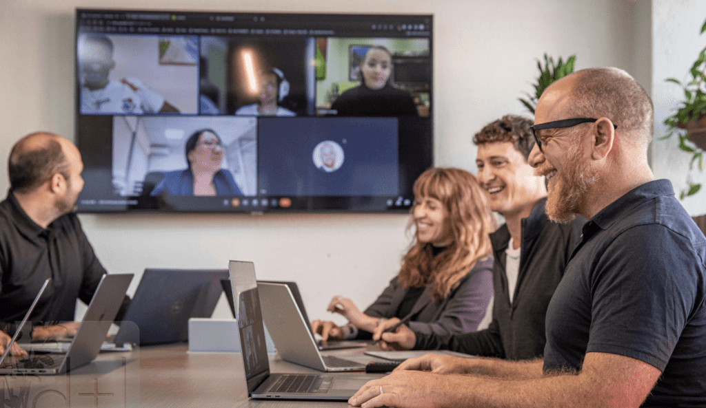 A smiling software development team with a client in profile, with a fourth person on a Zoom call in the background.