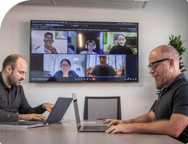 Two men on laptops with a group of five people attending the meeting virtually in the background