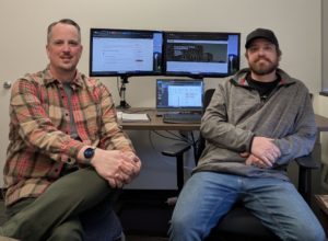 Two men seated in an office, each with dual monitors and a laptop displaying a lesson-sharing platform.